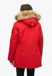 Parka roja con capucha forrada de piel sintética, que presenta una cintura ajustada y mangas largas. Incluye una insignia en la manga izquierda y una textura suave.
