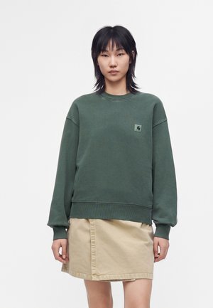 Jeune femme portant un sweat-shirt vert et une jupe beige, debout devant un fond clair uni, regardant devant avec une expression neutre.
