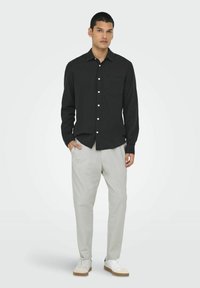 Camisa negra de botones con un bolsillo delantero, combinada con pantalones gris claro y zapatillas blancas. La camisa tiene mangas largas y un corte relajado.