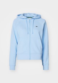 Sudadera con cremallera de color azul claro, confeccionada en tejido suave, que presenta un bolsillo tipo canguro, cordones ajustables y un pequeño logo en el pecho.