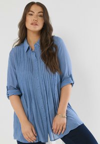 Donna con lunghi capelli castani mossi, che indossa una camicia azzurra chiara a maniche arrotolate e pantaloni scuri, in posa contro uno sfondo neutro.
