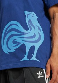 Blauwe katoen T-shirt met een groot gestileerd lichtblauw hanenlogo op de voorkant. Zwarte shorts met het witte Adidas-logo zichtbaar.