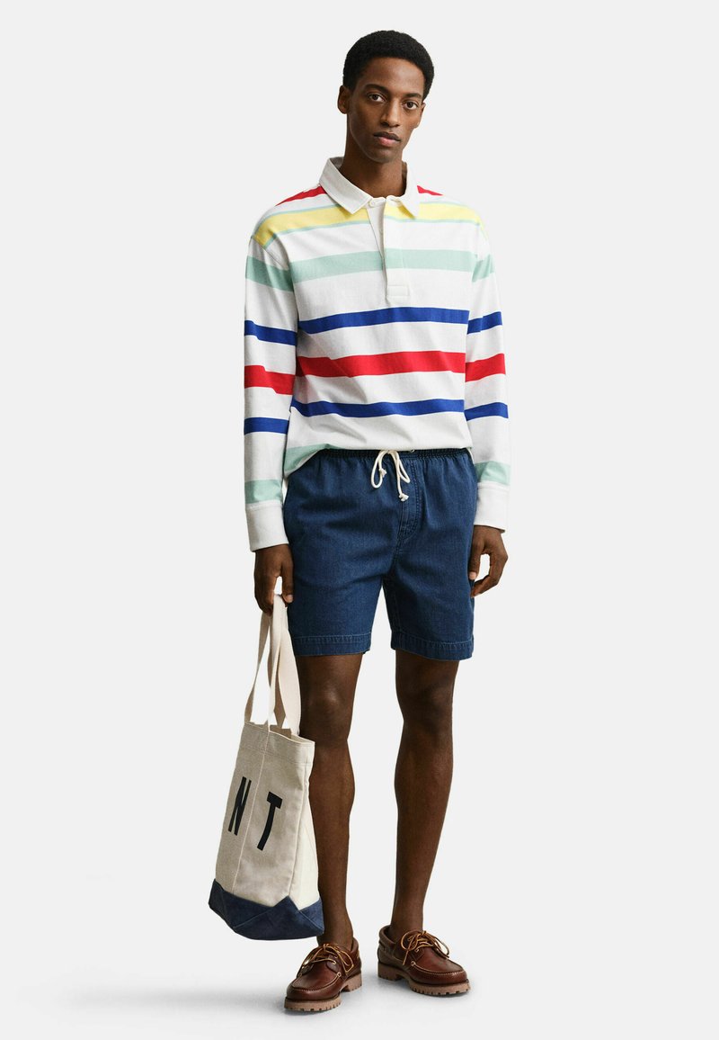 Giovane uomo in piedi che indossa un polo a maniche lunghe a strisce multicolori, pantaloncini blu navy, scarpe marroni e tiene una borsa a tracolla in tela.