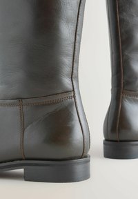 A barna bőr csizma sima felületű, kiemelkedő varrással és strapabíró fekete gumitalppal rendelkezik. A dizájn magas és kerek hátsó résszel készült.