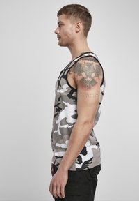 Pilkas ir baltas kamufliažinis tank top'as su juodu apvadu, pasižymintis laisvu pjūviu ir be rankovių dizainu. Matoma tatuiruotė ant viršutinio bicepso.