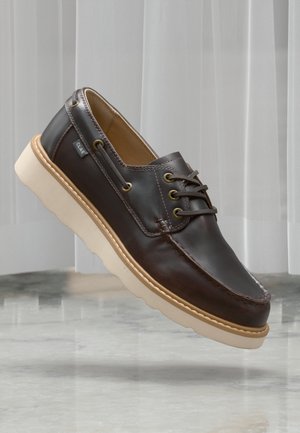 Zapato náutico de cuero marrón con suela de caucho cremosa y cordones en la parte delantera, exhibido sobre una superficie de mármol reflectante con un fondo de cortinas blancas translúcidas.