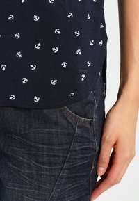 Mörkblå skjorta med vitt ankarmönster, tillverkad av lättviktig tyg. Rundad fåll, etikettdetalj på sidan, kombinerad med mörk denimjeans.