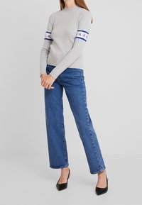 Maglione grigio a coste con collo alto e righe blu e bianche sulle maniche, abbinato a jeans a gamba larga blu e tacchi a punta neri.