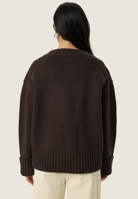 Pull en maille brun avec une coupe ample, des poignets et un ourlet côtelés, et un encolure large, présentant un motif texturé et un design à épaules tombantes.