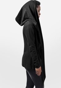 Sweat à capuche noir avec une capuche surdimensionnée et drapée, fabriqué en tissu doux. Présente une coupe longue et asymétrique ainsi qu'une poche avant. Porté avec un jean sombre.