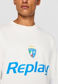 Hvid sweatshirt med blå tekst "Replay" og et farverigt skjoldlogo med tre stjerner ovenfor. 100% bomulds materiale, crew neck design.
