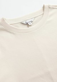 Šviesiai beige medvilninė marškinėliai su apvalia iškirpte, trumpomis rankovėmis ir etikete, nurodančia dydį S. Lygi tekstūra ir minimalus siuvimo detalumas.