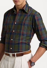 Camicia a quadri verde con bottoni, caratterizzata da linee blu, rosse e crema, con taschino sul petto e maniche arrotolate. Cintura e orologio in pelle marrone visibili.