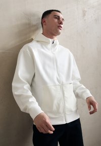 Sweat à capuche zippé blanc en tissu doux, avec une poche kangourou et des poignets côtelés. Texture lisse, design minimaliste et col à capuche.