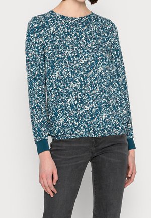 Kvinde iført en tealblå blomstret bluse med lange ærmer og ribkantede manchetter, kombineret med mørkegrå tætsiddende jeans, stående foran en ensfarvet baggrund.