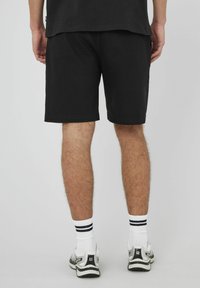 Shorts en coton noirs avec une coupe droite, ourlet fini, portés avec des chaussettes blanches rayées et des chaussures de sport grises, mettant en valeur un design simple.