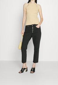 Beige tílko bez rukávů s výřezem na ramenou, spárované s černými kalhotami s vysokým pasem, které mají pět stříbrných knoflíků a kapsy. Černé skládací sandály.