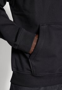 Sort sweatshirt med ribbede manchetter og kant, med en frontlomme og tekstureret stof. Materialet ser tykt og holdbart ud.