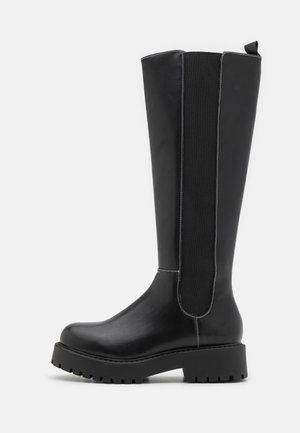 Botte noire en cuir montant jusqu'au genou avec semelle épaisse à crampons et panneau latéral élastique, présentant des détails de couture blanche et une languette de traction en haut.