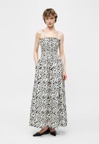 Femeie purtând o rochie albă fără mâneci, până la glezne, cu modele florale abstracte negre, bodice strâns pe talie, bretele subțiri și pantofi negri cu vârf ascuțit.