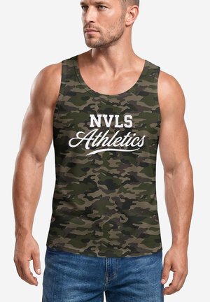 Muscular man wearing a green and brown camouflage tank top with "NVLS Athletics" text, paired with blue jeans, standing with arms relaxed.