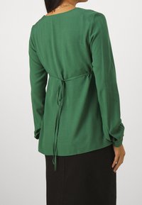 Kvinna klädd i en grön långärmad blus med en knytning i ryggen, tillsammans med en svart kjol, sedd från baksidan.