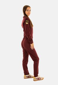 Femme en combinaison bordeaux avec des patchs de style militaire se tient de côté, portant des baskets beiges avec des rayures bordeaux sur fond blanc.