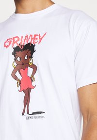 Camiseta de algodón blanca con un personaje de dibujos animados en un vestido rosa y pendientes de aro. El texto gráfico dice "GRIMEY" en rojo arriba.