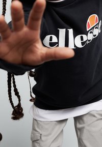 Felpa nera con logo "ellesse" bianco, caratterizzata da un emblema rotondo arancione e giallo. Sovrapposta a una t-shirt bianca, abbinata a pantaloni cargo beige chiaro.