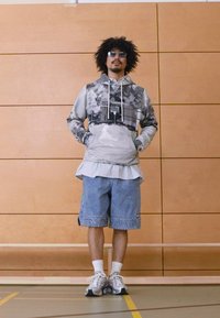 Šedá maskáčová mikina s kapucňou a šnúrkami, pod ňou biele tričko s pruhmi, rifľové šortky a tenisky. Doplnky zahŕňajú slnečné okuliare.