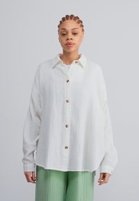 White, button-up shirt in textured fabric with a collar and brown buttons, paired with light green, pleated pants. Loose fit, long sleeves.