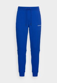 Niebieskie spodnie dresowe wykonane z miękkiego materiału, z elastycznym paskiem w talii i sznurkiem do regulacji, ściągaczami przy nogawkach oraz małym białym logo z boku.