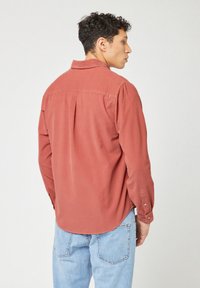 Roestkleurig lange mouwen shirt gemaakt van zachte stof, met een plooi op de rug, knoopmanchetten en een klassieke kraag. Draagt met lichtblauwe spijkerbroek.