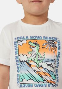 Witte katoenen t-shirt met een kleurrafisch van een groene krokodil die op golven surft, met palmbomen en de tekst "CALA NOVA BEACH".