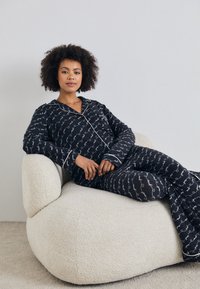 Svart pysjamassett med hvit bølgete mønster, med pipede kanter. Modell som sitter på en kremfarget, teksturert polstret stol.