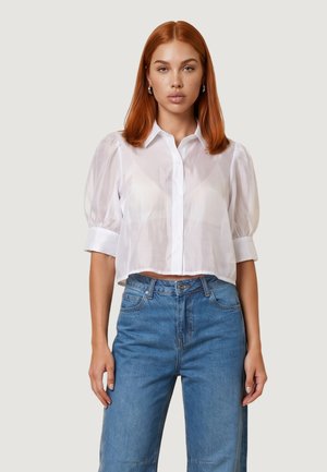 Femme aux cheveux roux portant un chemisier blanc transparent à manches bouffantes et un jean bleu taille haute, debout devant un fond clair uni.