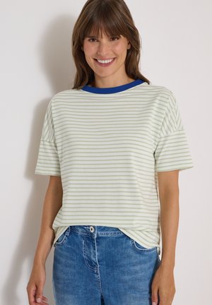 Femme souriante portant un t-shirt rayé vert clair et blanc avec un col bleu et une jupe en jean bleue, debout devant un mur clair uni.