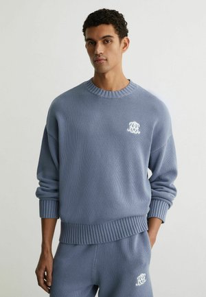Junger Mann trägt einen hellblauen Strickpullover und passende Hosen mit weißem Sticklogo auf Brust und Oberschenkel, steht vor schlichtem Hintergrund.