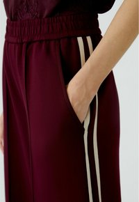Bordeauxfarbene Sporthose mit elastischem Bund, Seitentaschen und kontrastierenden cremefarbenen Streifen entlang der äußeren Nähte. Glatte Textur.