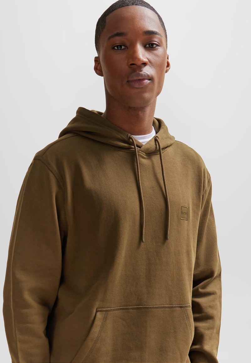 Brązowy sweatshirt z kapturem, z przednią kieszenią i sznurkiem. Posiada małą łatkę z logo na piersi. Wykonany z miękkiego materiału bawełnianego, luźny krój.