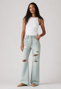 Femme aux longs cheveux bouclés portant un débardeur blanc, un jean large déchiré bleu clair et des chaussures plates beiges, debout devant un fond uni.