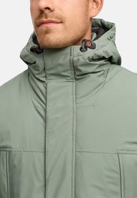 Uomo che indossa un giacca invernale verde salvia con colletto alto, dotata di cordini per cappuccio regolabili e toggles neri in plastica.