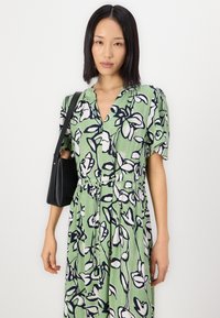 Grønn kjole med blomsterprint i svart og hvitt, med korte puffermer og V-hals. Modell med en liten svart håndveske.