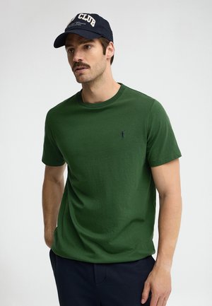 Uomo con t-shirt verde scuro a maniche corte, pantaloni blu navy e cappellino da baseball blu navy con il testo "CLUB", in piedi davanti a uno sfondo chiaro e uniforme.
