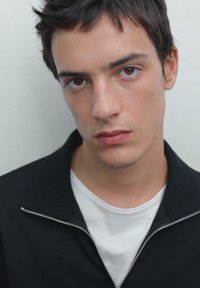 Zwarte zip-up jas over een wit T-shirt, close-up portret met kort haar en neutrale uitdrukking tegen een effen achtergrond.