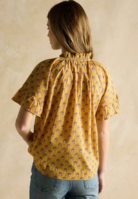 Kollane lillemustriga pluus, millel on kogutud kaelus ja lühikesed volangvarrukad, koos keskmise pesu denimist teksadega.