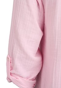 Ärmelsaum eines rosa strukturierten Hemdes, hochgekrempelt mit kleinem Knopfmanschetten, vor weißem Hintergrund.