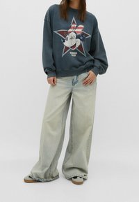 Donkergrijze sweatshirt met een afbeelding van Minnie Mouse tegen een sterrenachtergrond. Gecombineerd met lichtblauwe jeans met wijde pijpen. Bruine en grijze schoenen.
