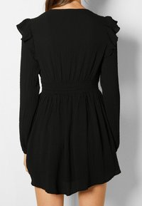 Schwarzes Kleid mit langen Ärmeln, strukturiertem Stoff, Rüschendetails an den Schultern, tailliertem Schnitt und ausgestellt Rock, von hinten gezeigt.