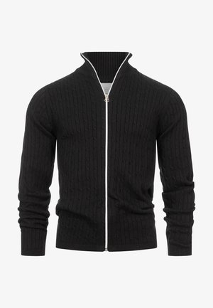 Čierny pletený sveter na zips s textúrovaným vrkočovým vzorom, so žebrovaným golierom a manžetami, v kombinácii s bielym tričkom a béžovými nohavicami.